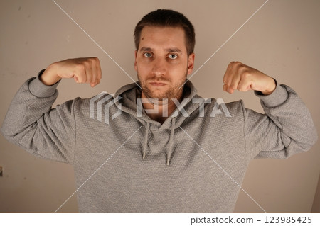 Flexing both arms, the man demonstrates his biceps and clenched fists. His pose emphasizes his physical strength and confidence. This gesture radiates power and pride in his physique. 123985425