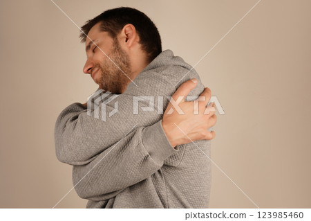 His expression suggests self-compassion and emotional connection. It s a soothing moment. The young man, with a light beard, embraces himself with both arms, exuding a sense of warmth and comfort. His expression suggests self-compassion and emotional connection. It s a soothing moment. The young man, with a light beard, embraces himself with both arms, exuding a sense of warmth and comfort. 123985460