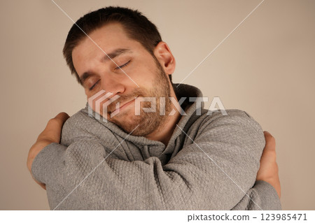 With a light beard, the young man holds himself close, offering a sense of warmth and serenity. His embrace highlights emotional comfort and self-affection. It s a tender, cozy moment. With a light beard, the young man holds himself close, offering a sense of warmth and serenity. His embrace highlights emotional comfort and self-affection. It s a tender, cozy moment. 123985471