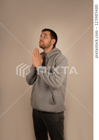 The pose expresses sincerity and devotion. In sporty attire, the young man clasps his hands together, creating a prayer-like gesture. His eyes are focused, suggesting a moment of hope. 123985480