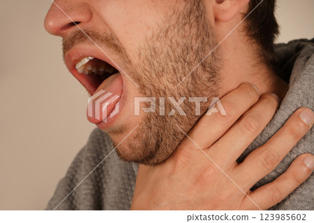 His facial expression conveys discomfort and a possible choking sensation. A man holds his throat with one hand, opening his mouth as if he is coughing or struggling to breathe. 123985602
