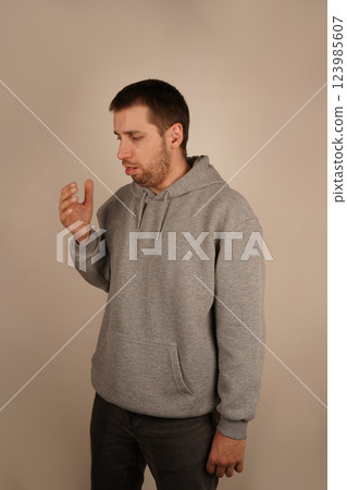 Young man in a gray hoodie standing indoors. He appears to be engaged in thought or mid-gesture, creating a natural and casual moment. Neutral background highlights the simplicity of the scene. 123985607
