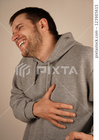 A man in a gray hoodie holds his chest near the ribs with one hand, his face showing pain. The expression suggests a burning sensation or severe discomfort. It could indicate heart failure symptoms. 123985615