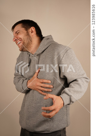 It could indicate heart failure symptoms. A man in a gray hoodie holds his chest near the ribs with one hand, his face showing pain. The expression suggests a burning sensation or severe discomfort. It could indicate heart failure symptoms. A man in a gray hoodie holds his chest near the ribs with one hand, his face showing pain. The expression suggests a burning sensation or severe discomfort. 123985616