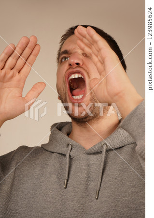 His actions suggest a loud announcement. The image conveys excitement and energy. With dark hair and a lively expression, the man cups both hands over his mouth, resembling a megaphone. 123985624