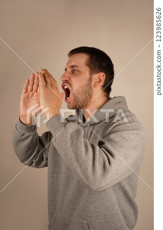 A young man with dark hair places his hands near his mouth, resembling a megaphone. His enthusiastic pose suggests he is delivering an energetic shout. The scene captures his dramatic gesture. 123985626