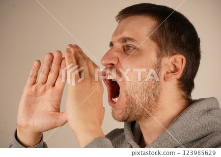 His enthusiastic pose suggests he is delivering an energetic shout. The scene captures his dramatic gesture. A young man with dark hair places his hands near his mouth, resembling a megaphone. His enthusiastic pose suggests he is delivering an energetic shout. The scene captures his dramatic gesture. A young man with dark hair places his hands near his mouth, resembling a megaphone. 123985629