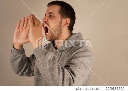 His actions suggest a loud announcement. The image conveys excitement and energy. With dark hair and a lively expression, the man cups both hands over his mouth, resembling a megaphone. His actions suggest a loud announcement. The image conveys excitement and energy. With dark hair and a lively expression, the man cups both hands over his mouth, resembling a megaphone. 123985631