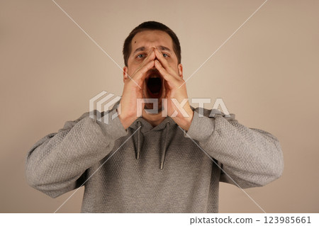 His animated gesture implies shouting or announcing something important. His expression radiates urgency. The dark-haired man holds both palms around his mouth, forming a makeshift megaphone. His animated gesture implies shouting or announcing something important. His expression radiates urgency. The dark-haired man holds both palms around his mouth, forming a makeshift megaphone. 123985661