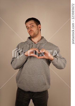His expression radiates kindness and warmth. A young man with light stubble makes a heart gesture with his fingers. Wearing a gray hoodie, he stands against a neutral background. His expression radiates kindness and warmth. A young man with light stubble makes a heart gesture with his fingers. Wearing a gray hoodie, he stands against a neutral background. 123985672