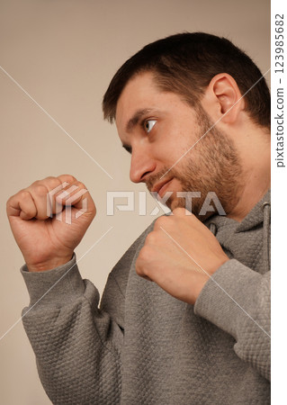 His posture suggests readiness and determination. The image conveys a strong and focused attitude. A man in a sporty hoodie stands with clenched fists in a boxing stance. His posture suggests readiness and determination. The image conveys a strong and focused attitude. A man in a sporty hoodie stands with clenched fists in a boxing stance. 123985682