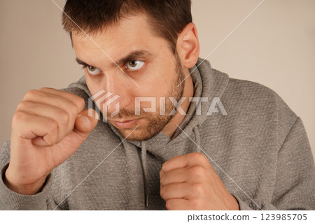 The scene suggests physical preparation and mental resilience. The man in an athletic hoodie adopts a boxing pose with tightly clenched fists. His stance exudes strength and focus. The scene suggests physical preparation and mental resilience. The man in an athletic hoodie adopts a boxing pose with tightly clenched fists. His stance exudes strength and focus. 123985705