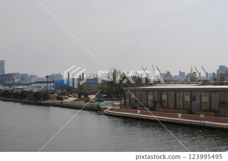 A view of a streetscape with a pale blue sky, the sea, warehouses on the opposite bank, and various other things. 123995495