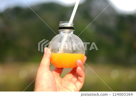 Closed up Asian woman hand holding fresh orange juice in clear glass bottle at outdoor with mountain in natural sunlight 124003164