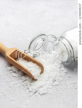 Magnesium chloride flakes being poured out of a jar onto a gray table with wooden scoop 124004637