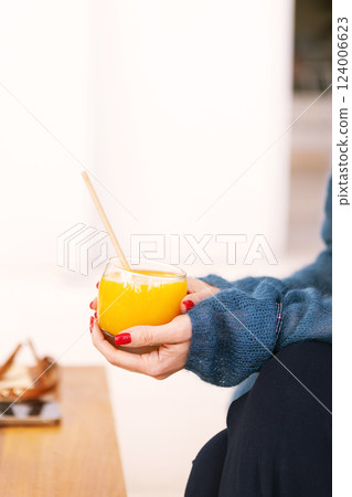 Close-up hands of woman holding glass of fresh orange juice, refreshing citrus drink for healthy lifestyle, minimalistic composition with vibrant blue and orange color and summer vibes 124006623
