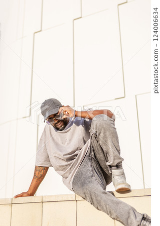 Handsome Black man in stylish streetwear sitting outdoors in city against white wall, wearing gray t-shirt, cap, sunglasses, and jeans on sunny day 124006634