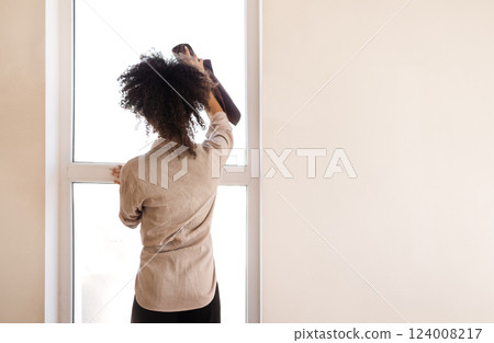 A curly-haired mixed-race girl in casual clothes wipes the window with a rag. A young African woman cleans a house or apartment. 124008217