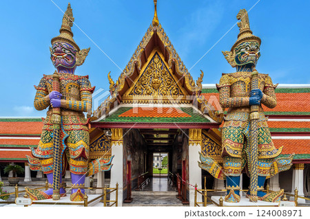 Two giant Yaksha guardian statues stand majestically at the entrance of the magnificent Wat Phra Kaew, adorned with intricate golden details and a striking multi-colored roof beneath a clear blue sky. 124008971