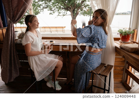 Girls Lakeside Cafe Summer: Two girls enjoy drinks at a lakeside cafe during summer, taking photos and relaxing. 124009448