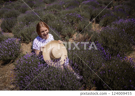 A woman is sitting in a field of lavender flowers and holding a straw hat A woman is sitting in a field of lavender flowers and holding a straw hat 124009455
