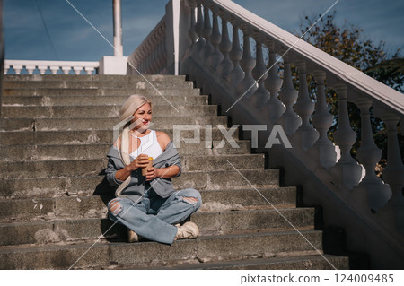 A woman sits on a set of stairs, holding a cup of coffee. She is enjoying her coffee and taking in the view from the top of the stairs. 124009485