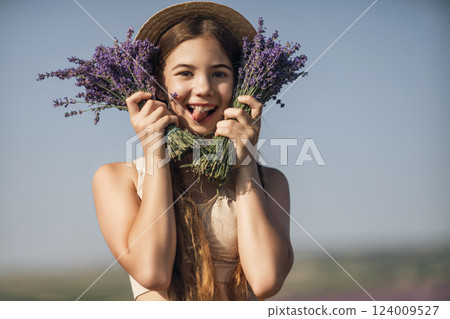 girl is holding a bunch of lavender purple flowers in her hands and wearing a straw hat. She is smiling and she is enjoying herself. The scene is set in a field of lavender, which adds to the peaceful 124009527