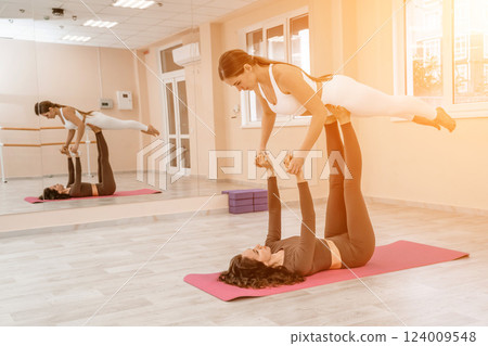 Two women do acroyoga on a light floor near a large mirror in a yoga studio. They are wearing leggings and tops. Two women do acroyoga on a light floor near a large mirror in a yoga studio. They are wearing leggings and tops. 124009548