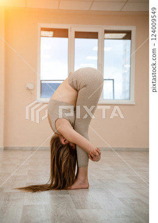 Young woman doing yoga in the gym. A girl with a long braid and in a beige tracksuit stands in a stork pose on a pink carpet. A woman performs padahstasana. Young woman doing yoga in the gym. A girl with a long braid and in a beige tracksuit stands in a stork pose on a pink carpet. A woman performs padahstasana. 124009549