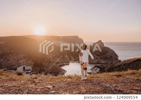 A woman stands on a hill overlooking a body of water. The sun is setting in the background, casting a warm glow over the scene. The woman is enjoying the view and taking in the beauty of the landscape A woman stands on a hill overlooking a body of water. The sun is setting in the background, casting a warm glow over the scene. The woman is enjoying the view and taking in the beauty of the landscape 124009554