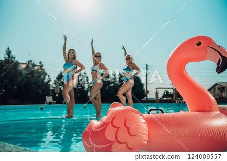 Three women are in the water, one of them is holding a pink flamingo. Scene is fun and playful. 124009557