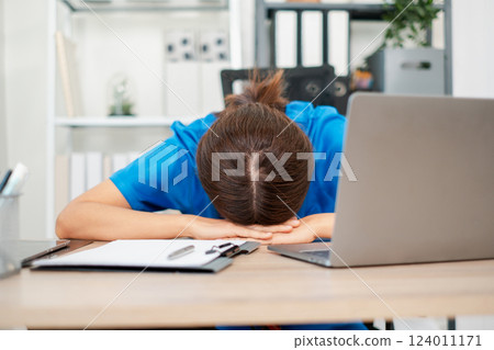 Tired healthcare professional in blue scrubs resting head on desk, surrounded by laptop and paperwork in a contemporary office setting. Tired healthcare professional in blue scrubs resting head on desk, surrounded by laptop and paperwork in a contemporary office setting. 124011171