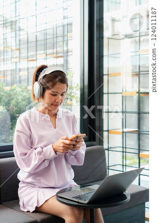 A young woman wearing headphones, using a smartphone in a modern cafe, with a laptop on the table. 124011187