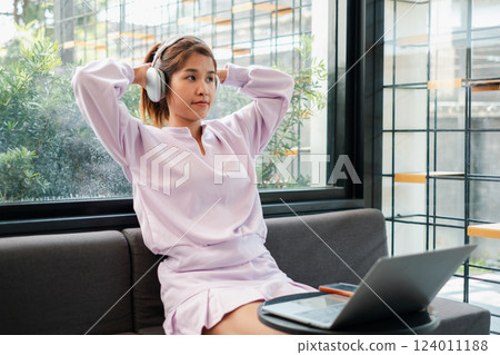 Woman wearing headphones, sitting on a sofa with a laptop, enjoying music in a modern cafe. 124011188
