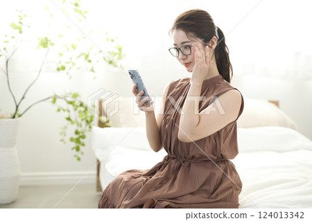 年輕女子坐在床上，看著她的智能手機 124013342