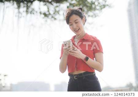Asian cheerful woman in red shirt and black jeans smiles while using her smartphone outdoors on sunny day Asian cheerful woman in red shirt and black jeans smiles while using her smartphone outdoors on sunny day 124014373