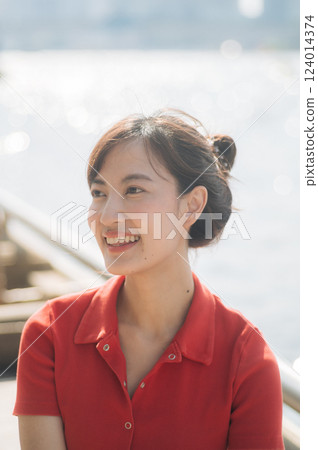 Asian cheerful woman in red shirt smiles brightly while sitting outdoors near waterfront on sunny day 124014374