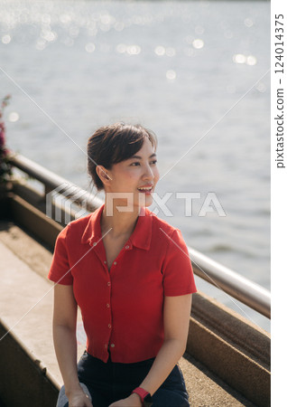 Asian cheerful woman in red shirt sits by waterfront, enjoying sunny day with serene expression Asian cheerful woman in red shirt sits by waterfront, enjoying sunny day with serene expression 124014375