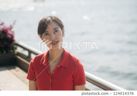 Asian woman in red shirt stands outdoors near waterfront, with soft sunlight and serene background 124014376