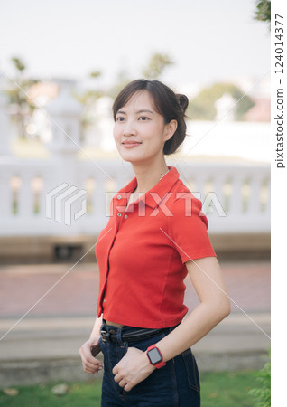 Asian young woman wearing red shirt and smartwatch stands outdoors, smiling confidently in park setting with blurred background Asian young woman wearing red shirt and smartwatch stands outdoors, smiling confidently in park setting with blurred background 124014377