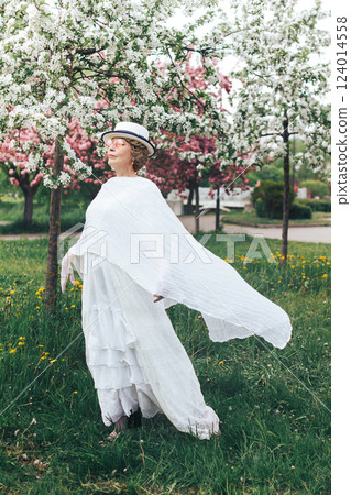 Happy natural adult mature woman of 60 years old in 18th century style white canotier hat, retro white dress, gloves and glasses in blossoming white and pink apple orchard on summer day 124014558