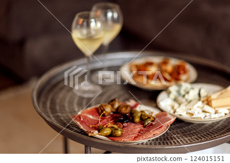 Two glasses of cool champagne and plates with various snacks on a round metal coffee table. White plates with slices of ham, salami, capers, olives and cheese. 124015151