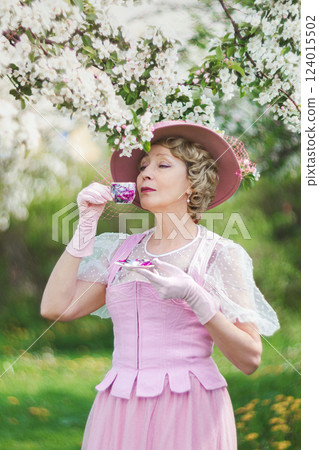 Happy natural adult mature woman of 60 years old in 18th century style pink hat, retro dress, gloves drinks tea in blossoming white apple orchard on summer day 124015502