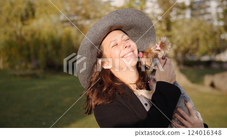In a vibrant and sunny park setting, a cheerful woman wearing a stylish hat playfully hugs her adorable dog In a vibrant and sunny park setting, a cheerful woman wearing a stylish hat playfully hugs her adorable dog 124016348