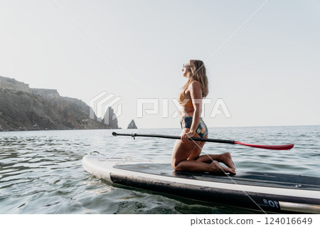 Woman Paddleboarding Ocean Coastline - Young woman kneels on paddleboard in calm ocean waters with rocky coastline in background. Woman Paddleboarding Ocean Coastline - Young woman kneels on paddleboard in calm ocean waters with rocky coastline in background. 124016649