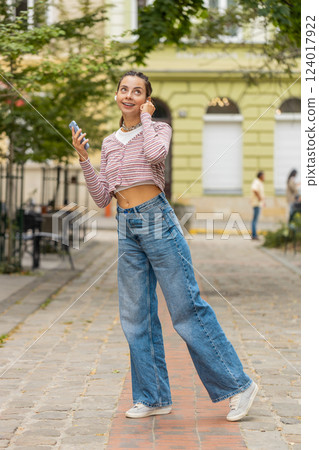 Happy relaxed woman standing on city street in wireless earphones listening dancing music outdoors Happy relaxed woman standing on city street in wireless earphones listening dancing music outdoors 124017922