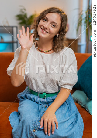 Happy young woman vlogger smiling friendly at camera and waving hands while sitting on sofa at home 124017928