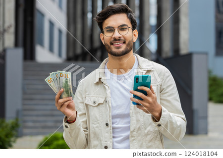 Happy Indian man counting money dollar cash, use smartphone calculator app in urban city street 124018104