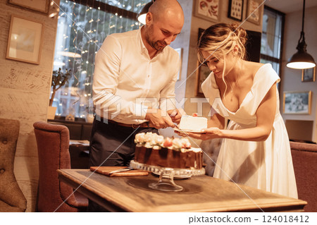 Attractive groom and charming bride are cutting festive wedding cake. Beautiful girl in white dress holds plate. Young man puts piece of sweetness on plate. Attractive groom and charming bride are cutting festive wedding cake. Beautiful girl in white dress holds plate. Young man puts piece of sweetness on plate. 124018412