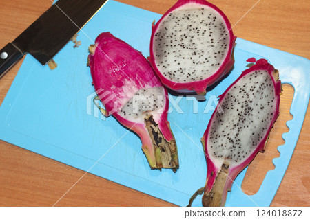 dragon fruit, sliced on blue plastic cutting board. half, quarter, pieces of tropical exotic fruit with bright pink peel and gray pulp with small black seeds, next to a large carving knife dragon fruit, sliced on blue plastic cutting board. half, quarter, pieces of tropical exotic fruit with bright pink peel and gray pulp with small black seeds, next to a large carving knife 124018872